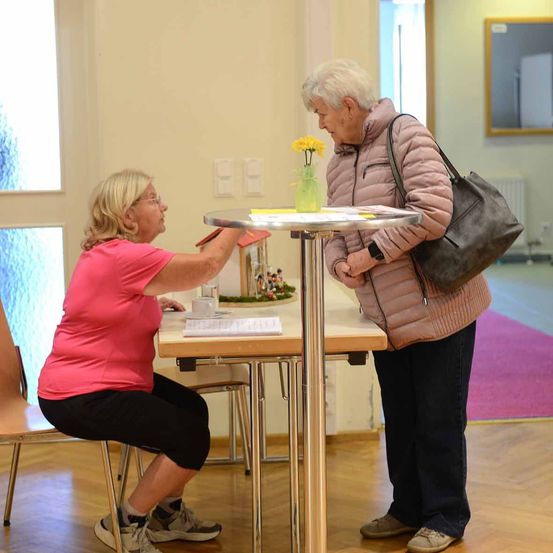 Zwei ältere Frauen stehen vor einem Tisch, eine hält eine Karte und die andere schaut sie an. Eine gelbe Blume steht in einer Vase auf dem Tisch. Der Raum hat einen Holzboden und weiße Wände.