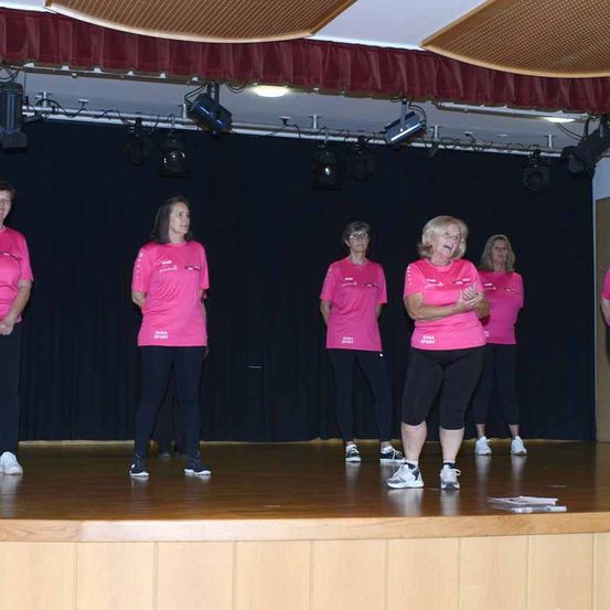 Fünf Frauen in rosa Shirts und schwarzen Hosen stehen mit verschränkten Händen auf einer Bühne. Sie scheinen an einem Fitness- oder Tanzkurs teilzunehmen.