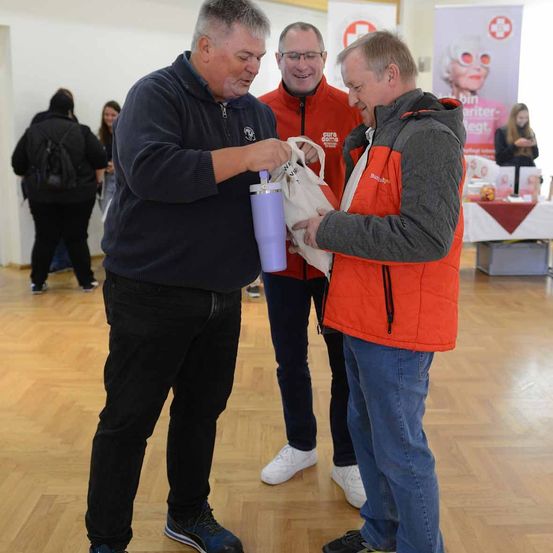 Drei Männer stehen auf einem Holzboden in einem Raum. Der Mann in der Mitte hält eine weiße Tasche, während der Mann auf der linken Seite einen lilafarbenen Becher hält. Hinter ihnen steht ein Mann in einem roten Mantel mit Brille. Es gibt andere Leute im Raum, einschließlich einer Frau mit einer Rucksack.