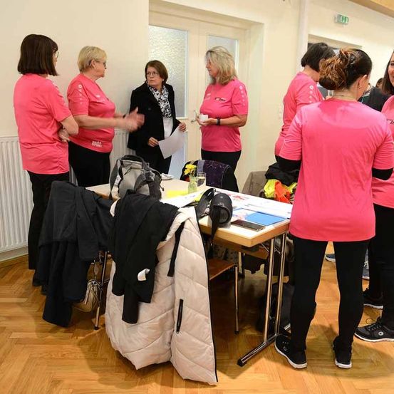 Eine Gruppe von Frauen in rosa Shirts und schwarzen Hosen steht in einem Raum, vielleicht bei einem Treffen. Vor ihnen steht ein Tisch mit Papieren, einer Tasche und einer gelben Blume.