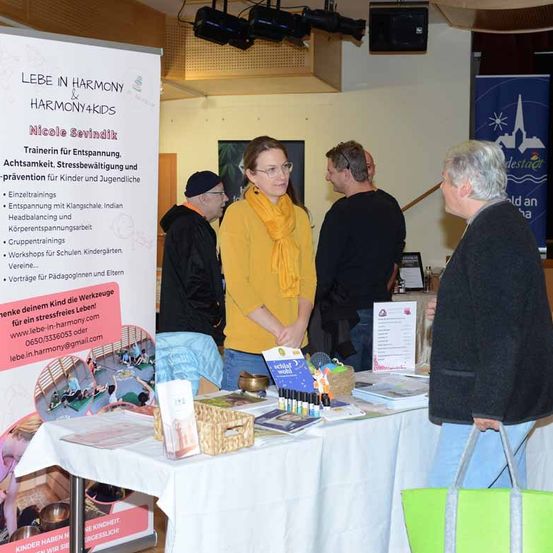 Eine Gruppe von Erwachsenen steht vor einem Tisch mit Materialien. Ein Banner im Hintergrund lautet 'Leben in Harmony & Harmony4Kids.' Sie diskutieren wahrscheinlich über Harmony4Kids, ein Programm für Stressmanagement und Prävention für Kinder und Jugendliche.