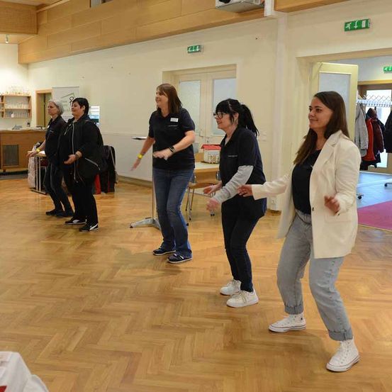 Fünf Frauen tanzen in einem Raum mit Holzböden. Drei von ihnen tragen eine Brille und zwei sind in Turnschuhen. Der Raum hat weiße Wände und Türen, mit einem Schreibtisch und Stühlen in der Ecke.