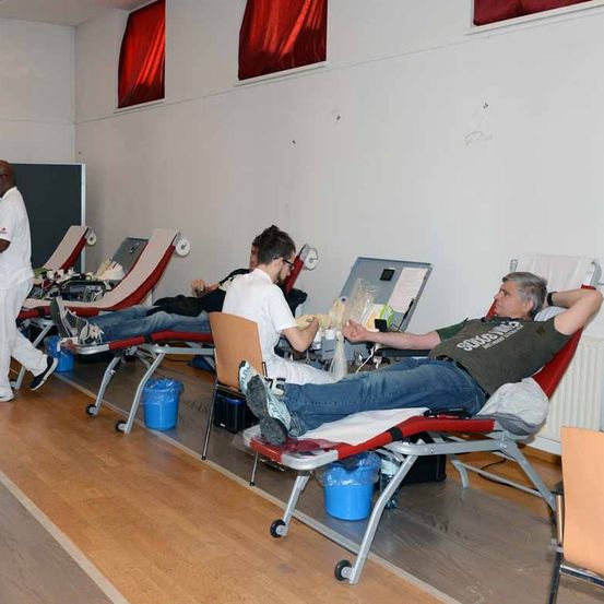 Ein Raum mit Holzböden und weißen Wänden. Mehrere Menschen liegen auf Betten, zwei erhalten Blutspenden. Ein medizinischer Mitarbeiter geht hinter den Betten.