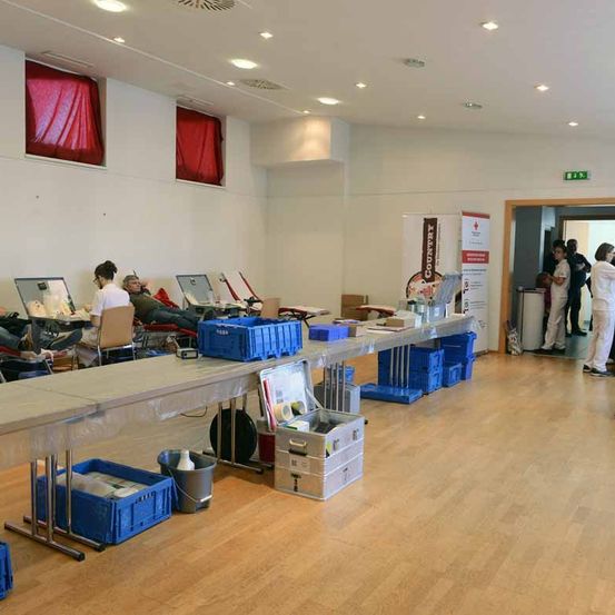 Ein Raum mit Holzböden und weißen Wänden hat mehrere stehen und sitzende Personen. Lange Tische mit Gegenständen und Kisten sind aufgestellt, eine Person sitzt in einem Stuhl. Ein Schild und eine Aufbewahrungsbox befinden sich rechts.