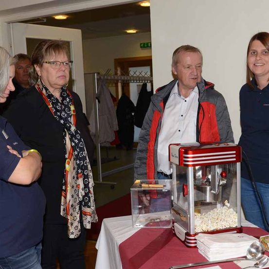 Vier Personen stehen um eine Popcorn-Maschine auf einem Tisch mit roter Tischdecke herum. Zwei Frauen und zwei Männer lächeln und schauen auf die Maschine.