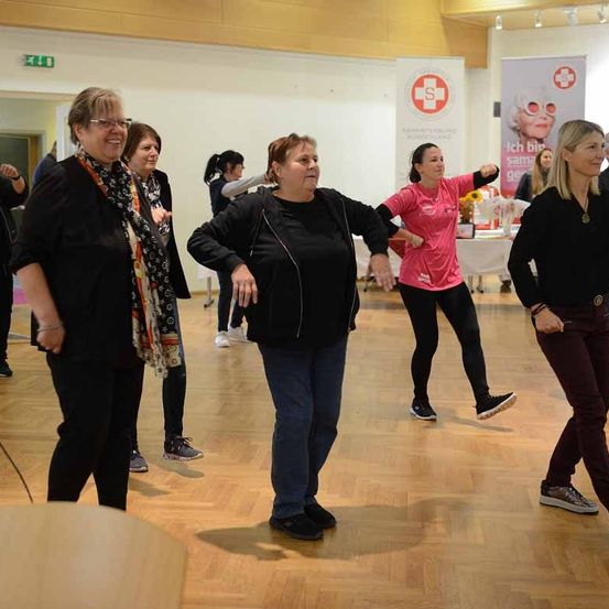 Eine Gruppe von Frauen tanzt in einem Raum. Die Frau in der Mitte trägt eine Jacke und lächelt. Die Frau auf der rechten Seite trägt ein rosa T-Shirt. Hinter ihnen steht eine Frau und lächelt. Im Hintergrund befinden sich Poster mit einer Frau und einem roten Kreuz.