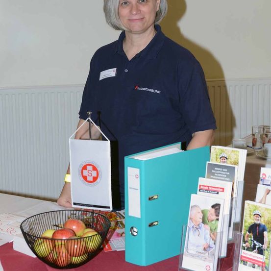 Eine Frau steht hinter einem Tisch mit einem Korb voller Äpfel, einer blauen Mappe und Broschüren. Sie trägt ein marineblaues Hemd mit dem Logo von Samariterbund.