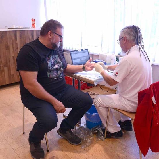 Ein Mann sitzt auf einem Stuhl und erhält von einem Mann in einem Laborkittel eine medizinische Behandlung. Ein Laptop steht auf dem Tisch zwischen ihnen, und ein Mantel hängt am Stuhl neben ihnen.
