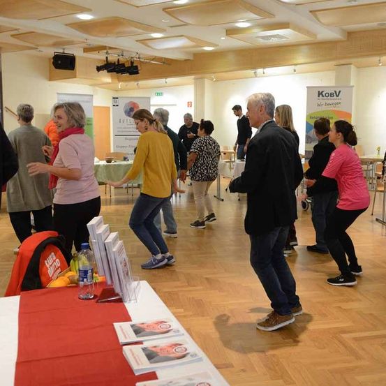 Eine Gruppe von Menschen tanzt in einem Saal mit Holzboden, einige tragen Turnschuhe. Ein Tisch mit Büchern und einer Flasche steht im Vordergrund. An den Wänden hängen Banner.
