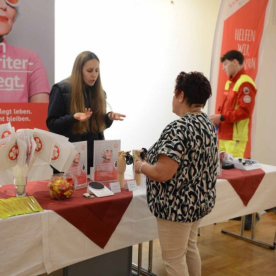 Zwei Frauen und ein Mann sind an einem Tisch. Eine Frau in einem Leopardenmusterhemd schaut sich eine Broschüre an. Eine andere Frau spricht und gestikuliert. Dahinter steht ein Mann in einem roten und gelben Mantel.