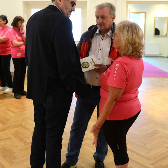 Ein Mann in einem schwarzen Anzug spricht mit einer Frau in einem rosa Shirt und hält ein Papier in der Hand. Hinter ihnen liest ein Mann in einem roten und schwarzen Jackett das Papier. Hinter dem Mann stehen zwei Frauen in rosa Shirts. Ein Spiegel ist an der Wand montiert.