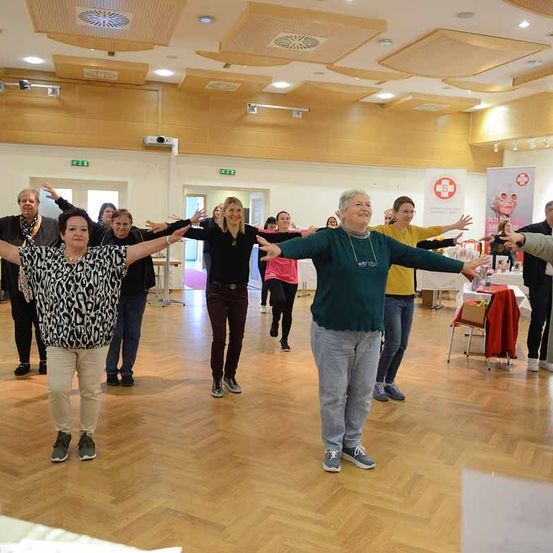 Eine Gruppe Erwachsener tanzt in einem Raum mit Holzboden. Sie tragen Freizeitkleidung. Ein Tisch mit rotem Tuch und einer weißen Fahne steht hinter ihnen.