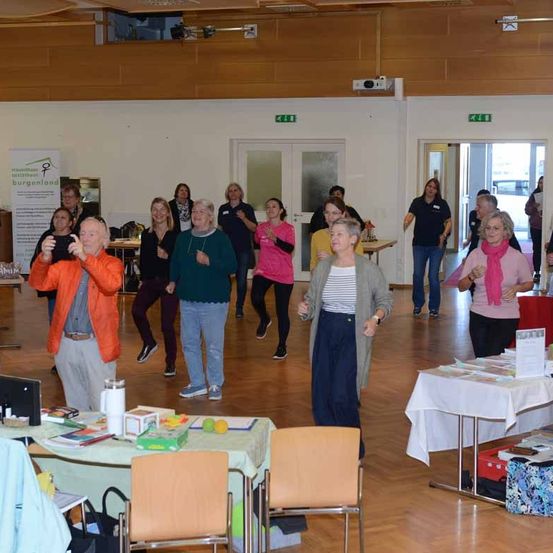 Eine Gruppe von Menschen befindet sich in einem Raum, wahrscheinlich an einer Veranstaltung teilnehmend. Ein Mann in Orange führt sie an, während andere zusehen. Tische, Stühle und Bücher sind in der Umgebung. Es gibt Türen und einen Projektor.