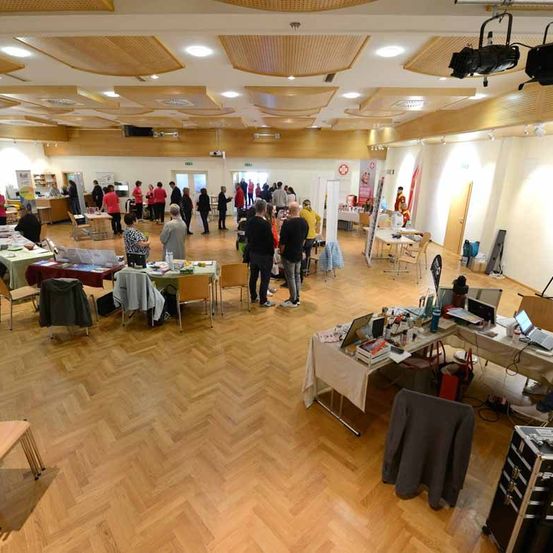 Ein großer Raum mit Holzböden und mehreren Tischen mit Gegenständen darauf. Es gibt Menschen, die herumlaufen, und eine Person in einem roten Shirt, die bei einem Tisch steht.