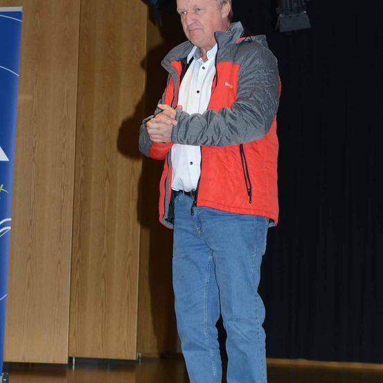 Ein Mann steht auf der Bühne, trägt eine rote und graue Jacke mit Kapuze, blaue Jeans und ein weißes Hemd. Er hält die Hände verschränkt und schaut nach vorne. Hinter ihm ist eine Holzwand und ein blauer Banner sichtbar.