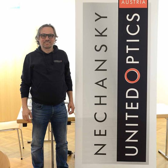 Ein Mann steht neben einem großen Banner mit dem Text 'Nechansky United Optics'. Er trägt eine Brille und eine schwarze Kapuzenjacke. Der Hintergrund ist ein Raum mit Holzböden und einem Schreibtisch.