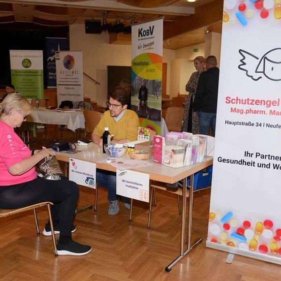 Eine Frau in einem rosa Shirt sitzt an einem Schreibtisch mit einem Mann in einem gelben Shirt. Sie sind von gesundheitsbezogenen Materialien und Bannern umgeben.