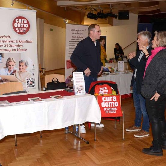 Mehrere Personen stehen um einen Tisch in einem Raum mit Holzboden. Auf dem Tisch befindet sich ein rotes Banner mit dem Text 'cura domo'. Ein Poster ist an der Wand hinter ihnen befestigt.