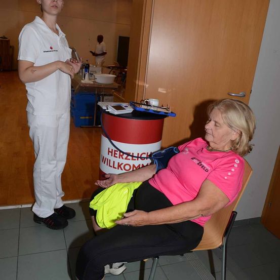 Eine Frau in einem rosa T-Shirt sitzt auf einem Stuhl und lässt ihren Blutdruck von einer Person in weißer Uniform überprüfen. Der Ort ist innen mit einem Holzboden.