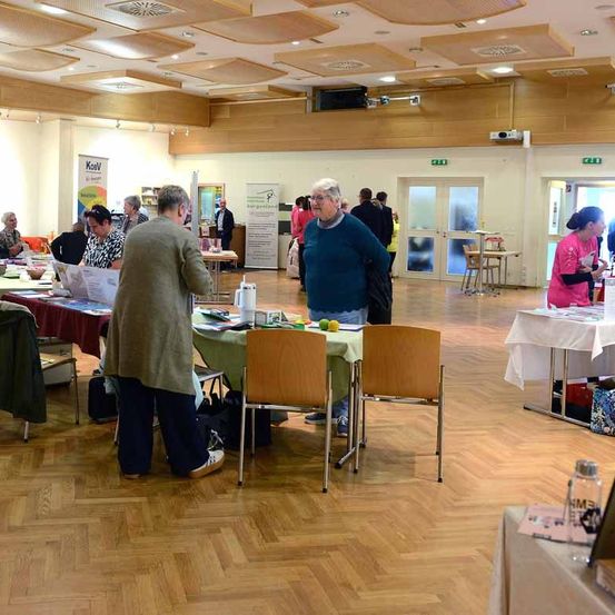 Eine Indoor-Veranstaltung mit mehreren Tischen, an denen Menschen Materialien durchblättern. Einige Tische haben weiße Tischdecken, andere grüne. Es gibt Stühle und ein Schild an der Wand.