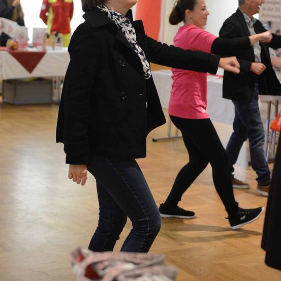 Eine Frau in einem schwarzen Mantel und einer Schärpe tanzt mit einem Mann und einer Frau in einem rosa Shirt in einem Raum mit Tischen und einer roten Tischdecke.