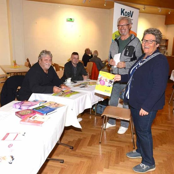 Eine Gruppe von Menschen ist in einem Raum, zwei stehen neben einem Tisch mit Büchern, während zwei andere dahinter sitzen. Eine Frau hält ein gelbes Buch und lächelt, und ein Mann steht mit einem Stück Papier.