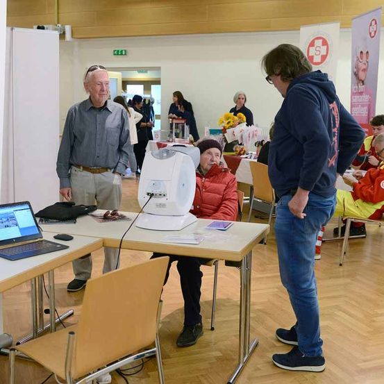 Ein älterer Mann steht neben einem Schreibtisch mit Laptop und einem Gerät, während ein anderer Mann ihn ansieht. Eine ältere Frau sitzt in einem Stuhl neben dem Schreibtisch. Andere Menschen sind anwesend, einige tragen gelbe Westen.