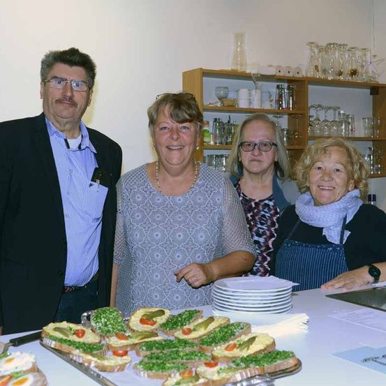 Vier Menschen stehen lächelnd hinter einem Tisch. Auf dem Tisch befinden sich verschiedene Sandwiches mit grünen Toppings. Ein Mann und eine Frau stehen an den Rändern, mit zwei Frauen dazwischen.