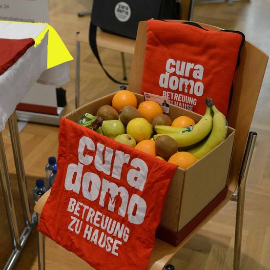 Ein Karton gefüllt mit verschiedenen Früchten wie Orangen, Bananen und Kiwis. Ein rotes Banner trägt die Aufschrift 'cura domo Betreuung zu Hause'. Ein weiterer roter Beutel liegt auf dem Stuhl hinter der Box. Ein gelber Aufkleber ist auf der linken Seite.