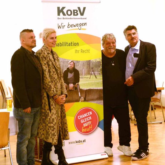 Vier Personen stehen vor einem Banner mit Text und Bildern, möglicherweise bei einer Veranstaltung. Das Banner hat die Worte 'Rehabilitation' und 'Ihr Recht'. Die Leute lächeln und scheinen für ein Foto zu posieren. Hinter ihnen steht ein Tisch mit einem Glas und einem Stuhl. In der Ferne sind auch Stühle und Tische.