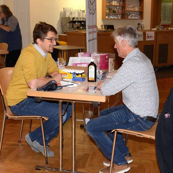 Zwei Männer sitzen an einem Tisch und unterhalten sich. Ein Mann lächelt, während der andere aufmerksam zuhört. Hinter ihnen ist eine Kaffeemaschine und ein Regal mit verschiedenen Gegenständen zu sehen.