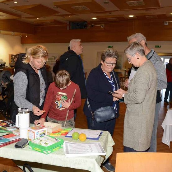 Eine Gruppe von Menschen ist um einen Tisch versammelt, auf dem verschiedene Gegenstände stehen, darunter Bücher, eine Zitrone und eine Kaffeetasse. Ein Junge hält einen Stock. Der Raum hat eine Holzdecke und ist gut beleuchtet.
