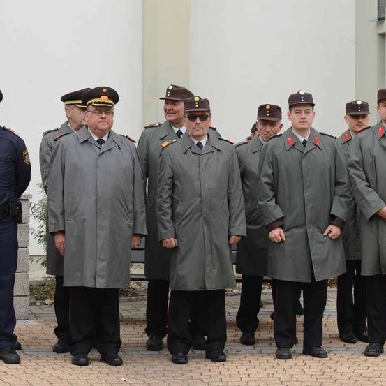 Eine Gruppe uniformierter Personen steht in Formation auf einem gepflasterten Gehweg. Sie tragen graue Mäntel und Mützen. Ein Mann in blauer Uniform steht abseits der Gruppe.