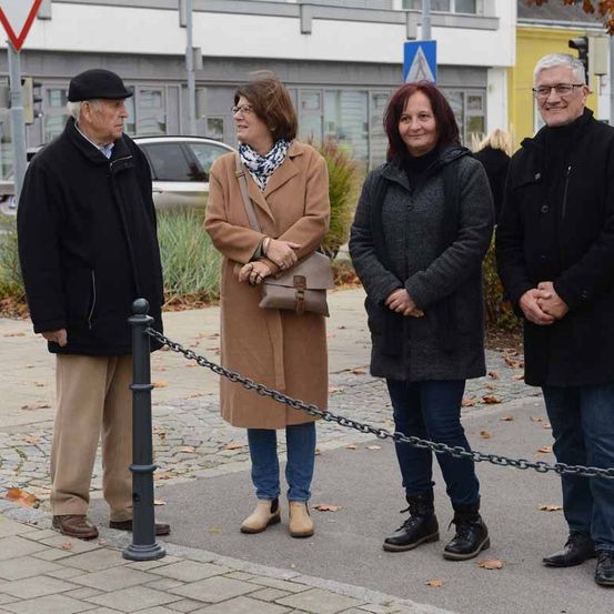 Vier Personen stehen in einem Park mit einem Kettengliederzaun, zwei Männer und zwei Frauen, wobei eine Frau eine Schürze und die andere Frau einen Mantel trägt. Hinter ihnen stehen Gebäude, ein Auto und ein Verkehrsschild.