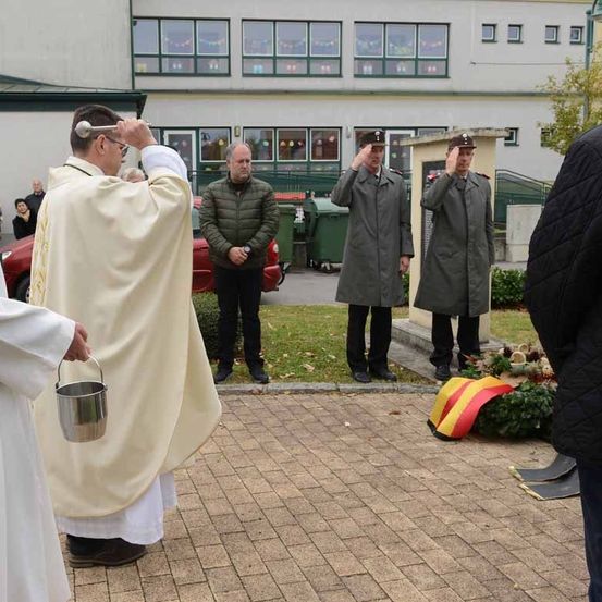 Ein Priester hält einen silbernen Eimer bei einer religiösen Zeremonie, wobei zwei uniformierte Männer salutieren und andere zuschauen. Hinter ihnen steht ein Gebäude mit Fenstern und einem roten Auto.