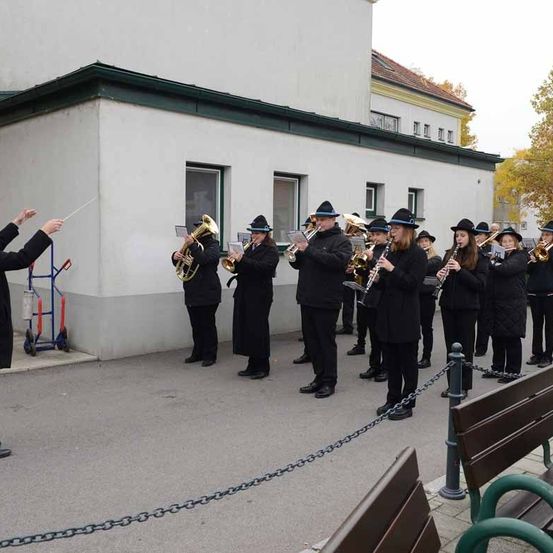 Eine Gruppe von Musikern in schwarzen Kleidungsstücken und Hüten spielt vor einem Gebäude, angeführt von einem Dirigenten mit einem Taktstock.