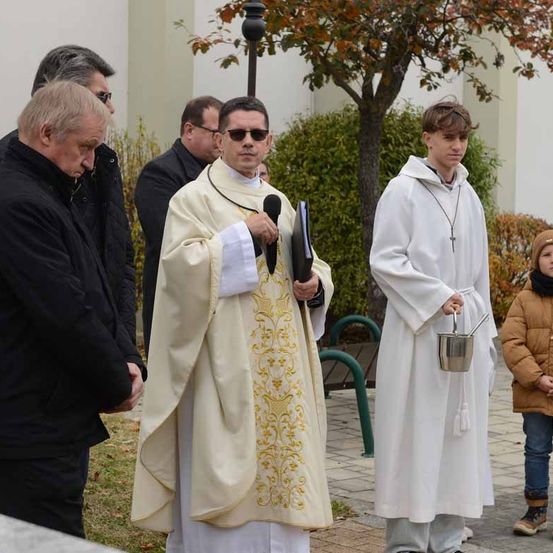 Ein Priester in zeremonieller Kleidung, der ein Buch und ein Mikrofon hält, steht neben einem Jungen, der einen silbernen Eimer hält. Andere Männer, darunter einer mit Brille, beobachten schweigend.