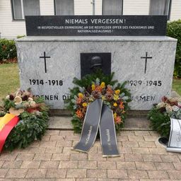 Ein Gedenkstein erinnert an die Opfer des Faschismus und Nationalsozialismus mit einem Kreuz, einem Porträthelm, Kränzen und Bändern. Die Daten 1914-1918 und 1939-1945 sind eingraviert.