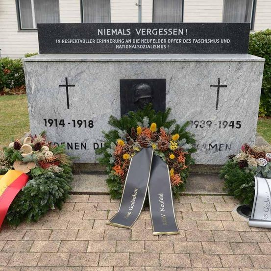 Ein Gedenkstein erinnert an die Opfer des Faschismus und Nationalsozialismus mit einem Kreuz, einem Porträthelm, Kränzen und Bändern. Die Daten 1914-1918 und 1939-1945 sind eingraviert.