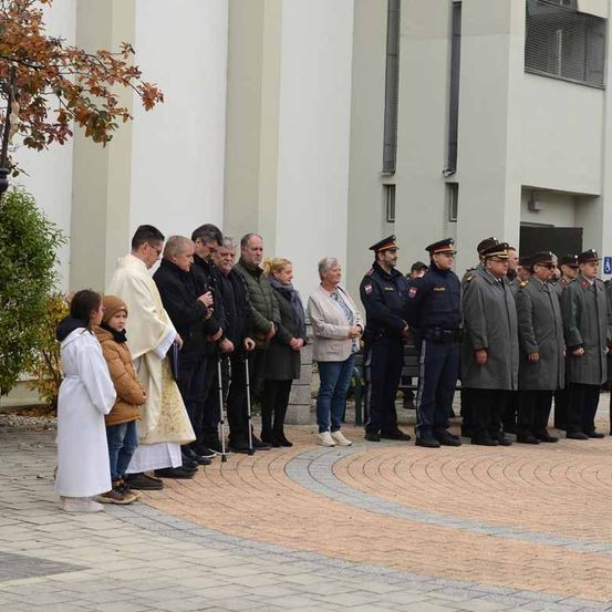 Eine Gruppe von Menschen in Uniform und Zivilkleidung steht im Kreis auf einem Ziegelboden. Ein Priester leitet ein Gebet mit einem kleinen Kind.