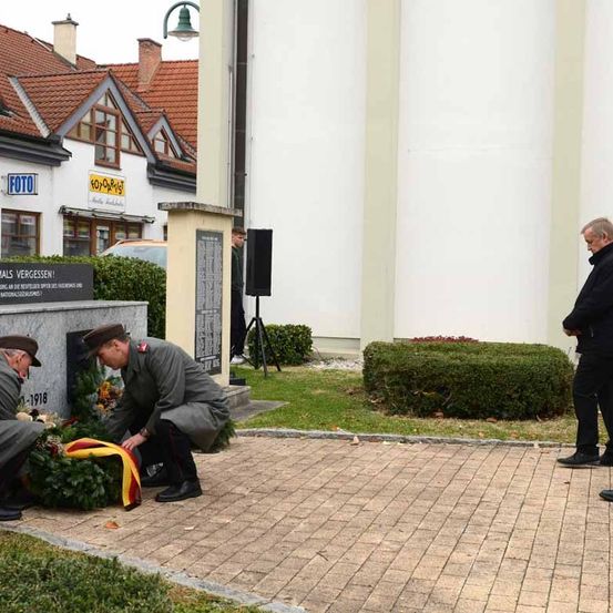 Zwei Männer in Uniform legen einen Kranz an einem Denkmal nieder. Ein Mann beobachtet von der Seite. Ein Gebäude mit einem Schild mit der Aufschrift Foto befindet sich hinter ihnen.