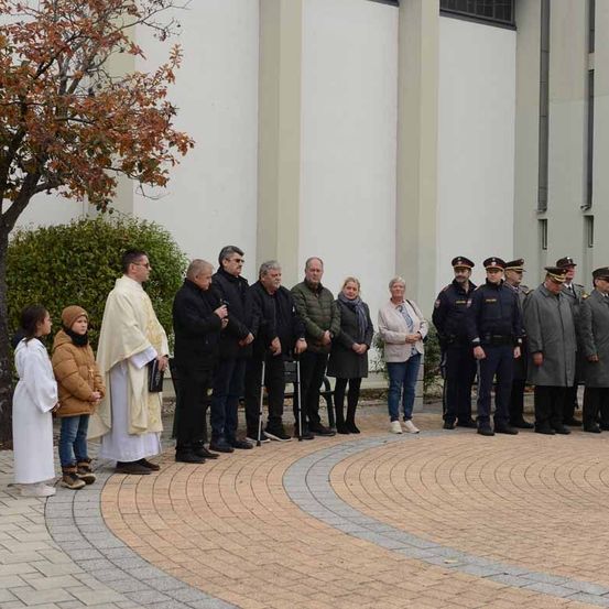 Eine Gruppe von Menschen, darunter ein Priester, steht im Freien in einem Kreis vor einem Gebäude. Einige tragen Mäntel, andere Uniformen. Ein Baum und Sträucher sind in der Nähe.