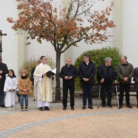 Ein Priester leitet eine religiöse Zeremonie vor einer Kirche, mit mehreren Personen, einschließlich Kindern, die in der Nähe stehen.