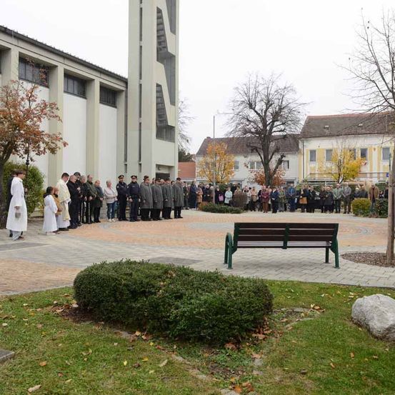 Eine Gruppe von Menschen steht auf einem Innenhof in der Nähe einer Kirche, mit uniformierten Beamten und Geistlichen vorne. Eine Bank und Sträucher sind in der Nähe, und Gebäude mit Bäumen im Hintergrund.