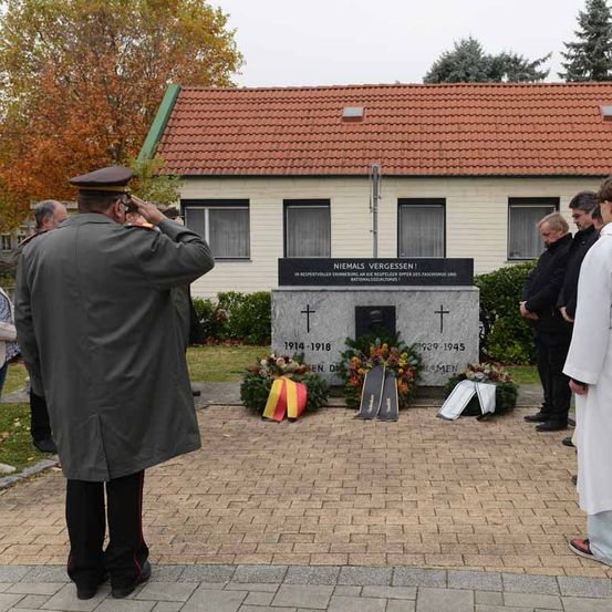 Eine Gedenkfeier findet an einem Denkmal statt. Ein Mann in Uniform salutiert, während andere hinter einer Girlande stehen. Das Denkmal hat zwei Kreuze und datiert von 1914 bis 1945.