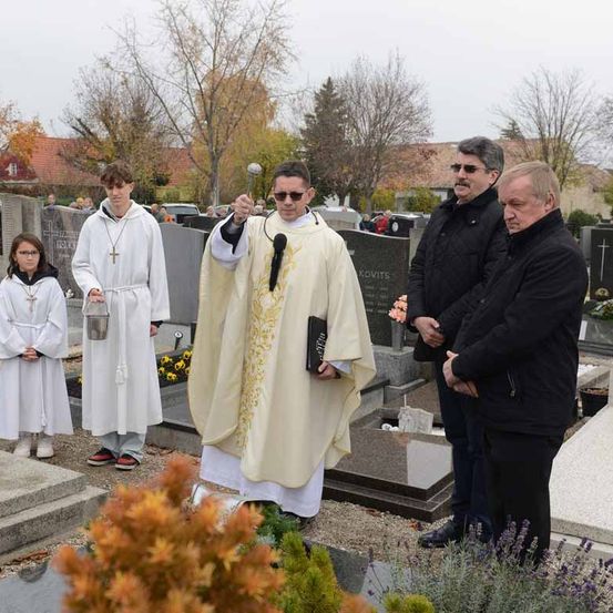 Ein Priester leitet eine Beerdigungszeremonie auf einem Friedhof, begleitet von zwei Messdienern und drei Männern in Winterkleidung.