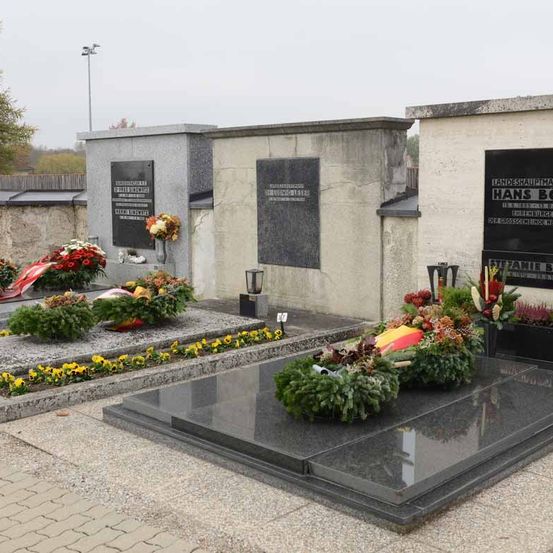 Ein Friedhof mit mehreren Grabsteinen, von denen einer für Hans Bog ist. Die Grabsteine haben Tafeln mit fremdsprachigem Text, und es gibt Blumen und Kränze um die Gräber.