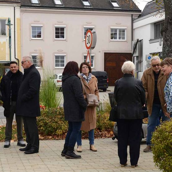 Eine Gruppe von Menschen steht auf einem Stadtplatz mit einem Geschwindigkeitsbegrenzungsschild im Hintergrund. Die Gruppe besteht aus Männern und Frauen, einige tragen Brillen und Mäntel.