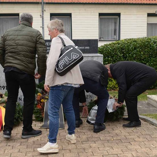 Vier Menschen stehen in der Nähe eines Denkmals, zwei beugen sich vor und legen Blumen nieder. Ein Mann in einer grünen Jacke und Jeans steht links. Das Denkmal hat Text und eine Girlande.