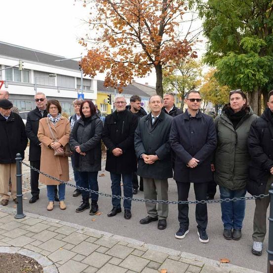 Eine Gruppe von Menschen in Winterkleidung steht in einer Reihe hinter einer Kettensperre auf einem gepflasterten Bereich. Dahinter befinden sich Gebäude, Bäume und Verkehrszeichen.